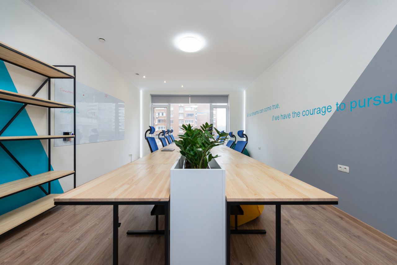 about-us Wooden table with green plants placed in light conference room with empty shelves and motivational inscription on wall in office building
